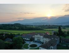 Veduta aerea di una grande villa storica con cortile centrale e edifici annessi dai tetti in coppi, immersa in campi e aree alberate. Sullo sfondo si estendono una pianura verde e una catena montuosa con il sole basso all’orizzonte.