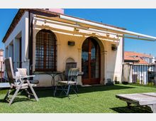 Terrazza sul tetto con prato sintetico, sedie e tavolino in legno, affacciata su un edificio con facciata chiara e grandi finestre ad arco con inferriate. Una tenda da sole si estende sopra l’ingresso e la ringhiera perimetrale guarda verso i tetti delle case vicine.