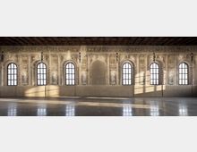 Ampia sala storica con soffitto a cassettoni in legno e lunga parete affrescata con finte architetture, nicchie e figure decorative tra grandi finestre ad arco. La luce naturale entra dalle finestre creando ombre sul pavimento liscio e lucido.