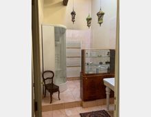 Bagno con pareti e pavimento in piastrelle chiare, doccia angolare con cabina curva in vetro e radiatore scaldasalviette a parete. Sono visibili una vetrina in legno con ripiani in vetro, un lavabo su colonna e tre lampade a sospensione decorative colorate dal soffitto.