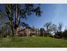 Grande villa in stile gotico con archi e torrette, situata su una collina erbosa in un parco. Intorno si vedono alberi alti, alcuni spogli e altri in fiore, sotto un cielo sereno.