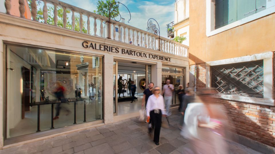 Ingresso di una galleria d’arte con ampia vetrata e portale centrale, insegna “Galeries Bartoux Experience” sopra la facciata chiara con balaustra in pietra. La scena è in una stretta calle lastricata tra edifici dai muri intonacati e in mattoni, con dettagli di finestre e grate.