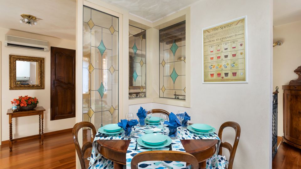 Sala da pranzo in un interno domestico con tavolo rotondo in legno apparecchiato, tovaglia a motivi blu e sedie coordinate su pavimento in parquet. Sullo sfondo si vedono pannelli vetrati decorati con motivi geometrici, una cornice con specchio, un piccolo tavolino con fiori e un quadro a parete.