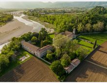 Veduta aerea di un complesso rurale storico con edifici in muratura e tetti in coppi rossi, immerso tra alberi e prati recintati. Sullo sfondo si vede un ampio fiume con greto ghiaioso, campi coltivati, un ponte ad arcate e una valle ai piedi di colline e montagne.