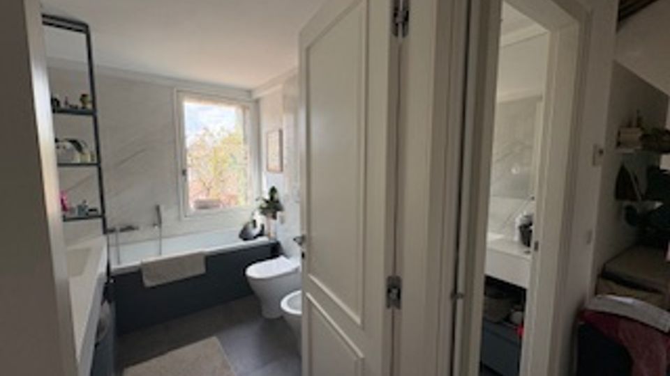 Bagno moderno e luminoso con vasca lungo la parete sinistra, lavandino e WC, piastrelle chiare e pavimento grigio. Una finestra sul fondo illumina la stanza e una porta aperta conduce a una camera da letto adiacente.