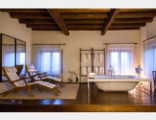 Interno di bagno in stile rustico con soffitto a travi in legno e pavimento in legno scuro, illuminato da grandi finestre con tende bianche. Al centro c’è una vasca freestanding con rubinetteria a vista; a lato due chaise longue con cuscini, specchi a parete e una struttura porta-asciugamani.