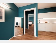 Interno di appartamento moderno con pareti verde petrolio e pavimento in legno, con un grande specchio a terra vicino a una porta che conduce alla camera da letto. A destra si vede una cucina lineare bianca con illuminazione sottopensile, lavabo e piano di lavoro.