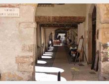 Portico in pietra con archi e travi in legno, che forma un passaggio coperto lungo una strada. Sul lato destro ci sono lavagne-menu e tavolini di un locale, con luce solare che crea chiazze sul pavimento in ciottoli.