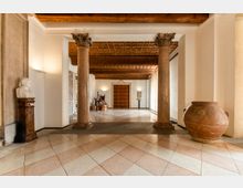 Interno di un atrio museale con pavimento a piastrelle geometriche, alto soffitto a cassettoni in legno e due colonne in pietra con capitelli decorati. Sulle pareti bianche sono esposti una scultura su piedistallo e un grande vaso in terracotta, con una porta in legno sul fondo.