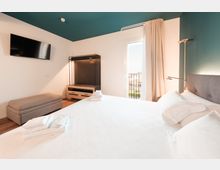 Camera da letto moderna con letto matrimoniale, pareti bianco e verde petrolio, TV a parete e mobile guardaroba aperto. Una portafinestra con tende chiare si apre su un piccolo balcone con ringhiera, illuminando la stanza con luce naturale.