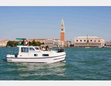Imbarcazione turistica bianca che naviga nella laguna, con sullo sfondo Piazza San Marco a Venezia, il campanile e il Palazzo Ducale affacciati sull’acqua sotto un cielo sereno.
