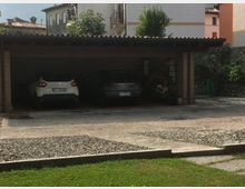 Garage coperto con tetto a spiovente in legno e pilastri in mattoni, contenente due automobili parcheggiate. Davanti al garage, vialetto in ghiaia e prato con erba; sullo sfondo, edifici residenziali e alberi.