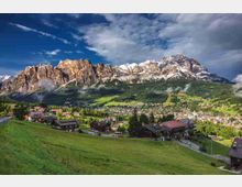 Panorama alpino con un paese tra prati verdi e boschi, visto da un pendio erboso in primo piano. Sullo sfondo si innalzano alte montagne rocciose con cime parzialmente innevate sotto un cielo nuvoloso.