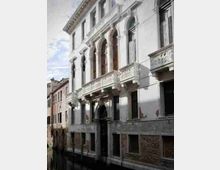 Facciata di un palazzo storico affacciato su un canale, con portale centrale ad arco e una serie di arcate al piano nobile. L’edificio presenta intonaco chiaro, finestre alte e balconi in pietra con balaustre scolpite, con edifici adiacenti visibili lungo la riva.