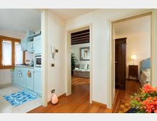 Interno di un appartamento con corridoio in parquet che collega una cucina compatta con mobili azzurri e finestra, un soggiorno con travi a vista e divano, e una camera da letto con armadio scuro e comodino con lampada. Pareti chiare e passaggi tra ambienti senza porte evidenti.