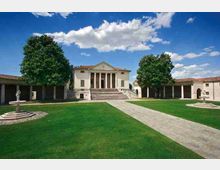 Cortile erboso di una villa in stile neoclassico con portico a colonne e scalinata centrale, affiancato da lunghe ali porticate. Un vialetto in pietra attraversa il prato verso l’edificio, con grandi alberi e due fontane ai lati sotto un cielo azzurro con nuvole.