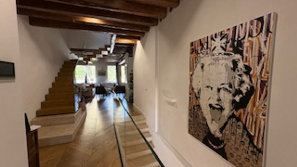 Interno di un’abitazione moderna con lungo corridoio in parquet, soffitto con travi in legno a vista e pareti bianche. A sinistra si vede una scala in legno e in fondo una zona giorno luminosa con grandi finestre; sulla parete destra è appeso un grande quadro in stile pop art.