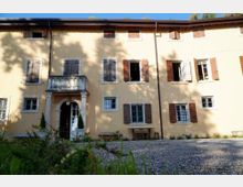 Facciata di una casa rurale a due piani intonacata in giallo, con finestre con scuri in legno e un ingresso ad arco con piccolo balcone soprastante. Davanti si vede un cortile in ghiaia con alcune piante e ombre di alberi proiettate sul muro.