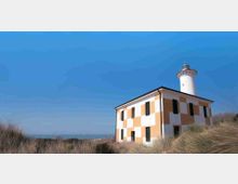 Faro costiero con torre cilindrica bianca e lanterna, collegato a un edificio rettangolare a due piani con facciata a riquadri bianchi e ocra e finestre scure. La struttura sorge tra dune con erba alta, con il mare all’orizzonte sotto un cielo azzurro.