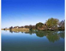 Paesaggio lacustre con acqua calma che riflette il cielo azzurro, canneti e una fascia di alberi lungo la riva. Sulla sponda si vedono piccole case in legno con tetto a spiovente e un pontile che si estende sull’acqua.