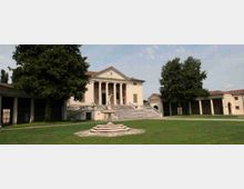 Villa in stile neoclassico con facciata a frontone e portico a colonne, raggiunta da un’ampia scalinata centrale. Davanti si estende un grande prato con una fontana al centro, alberi ai lati e porticati ad arcate che collegano gli edifici del complesso.