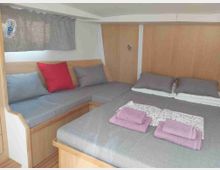 Cabina letto compatta con arredi in legno chiaro, grande letto con cuscini grigi e coperte piegate, e seduta angolare imbottita sotto una finestra con tenda. Pareti e soffitto bassi con piccoli dettagli integrati, tipici di un interno di barca o camper.