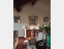 Soggiorno arredato in stile classico con soffitto a travi in legno, pareti chiare e pavimento in legno. In primo piano ci sono poltrone a righe e un tavolino, mentre sullo sfondo un mobile credenza con piatti decorativi e quadri appesi, accanto a un grande vaso con fiori.