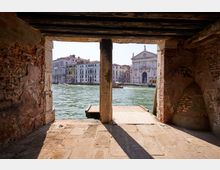 Vista da un portico in mattoni e pietra con travi in legno affacciato su un canale, con piccola banchina sull’acqua. Sullo sfondo si vedono palazzi storici lungo la riva e una chiesa con facciata classica in pietra chiara.