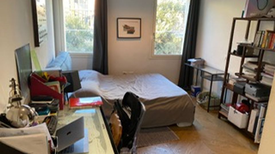 Camera da letto con scrivania e computer sulla sinistra, letto matrimoniale al centro e libreria metallica sulla destra. Due finestre con tende scure illuminano la stanza, con pavimento in parquet e lampada a sospensione.
