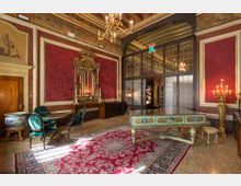 Salone interno in stile storico con pareti rosso scuro incorniciate, soffitto decorato e lampadari. Al centro ci sono un grande tappeto, un tavolo decorato e sedute, con una grande specchiera e un’ampia porta ad arco vetrata sul fondo.