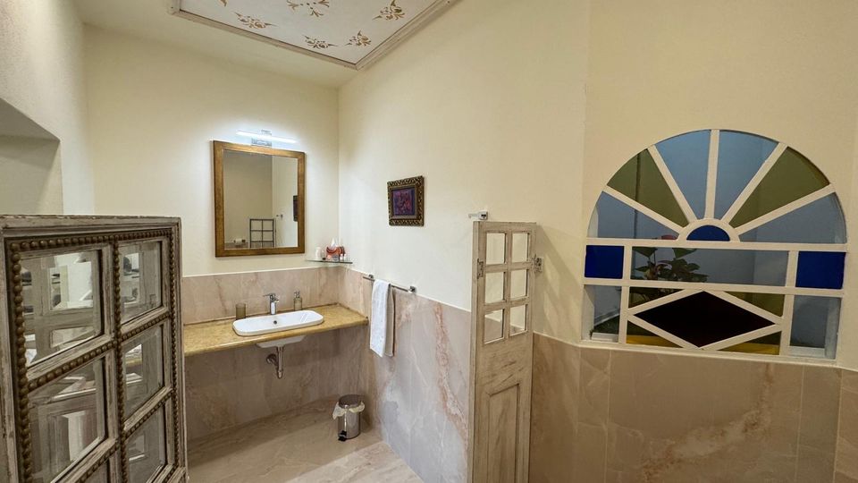 Bagno luminoso con pareti e pavimento in piastrelle chiare, lavandino con piano in pietra e grande specchio rettangolare sopra. A destra c’è una finestra ad arco con riquadri geometrici colorati e un soffitto alto con riquadro decorato.
