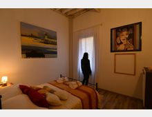 Camera da letto accogliente con letto matrimoniale in ferro battuto, copriletto a righe e asciugamani piegati; pareti con quadri e comodino con lampada. Al centro una finestra con tende bianche lascia filtrare luce naturale, con soffitto in travi a vista e pavimento in legno.