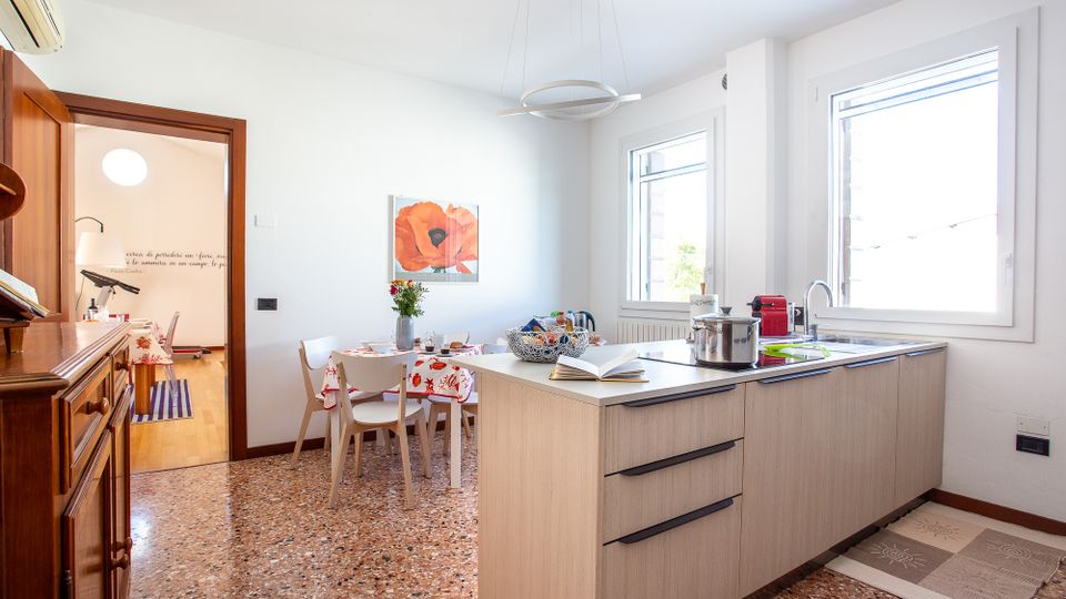 Cucina-soggiorno luminosa con isola moderna in legno chiaro e piano cottura, lavello sotto grandi finestre e pavimento in terrazzo. Sullo sfondo un tavolo da pranzo con sedie bianche, quadro a parete e passaggio verso un altro ambiente con pavimento in legno.