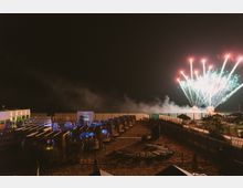 Spiaggia attrezzata di notte con file di cabine e pergolati illuminati, passerelle e recinzioni lungo la sabbia. Sullo sfondo, dal lato del mare, si vedono fuochi d’artificio che esplodono nel cielo sopra una fascia di fumo.