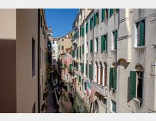 Canale stretto in una città storica, fiancheggiato da alti edifici in pietra con persiane verdi e finestre ad arco affacciate sull’acqua. Lungo le facciate sono appesi drappi e bandiere, e alcune gondole sono ormeggiate nel canale sotto un cielo sereno.