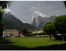 Ampio prato in una piazza cittadina con alberi potati in filare e edifici a due piani con tetti in tegole. Sullo sfondo si vedono montagne boscose e alte pareti rocciose sotto un cielo parzialmente nuvoloso.