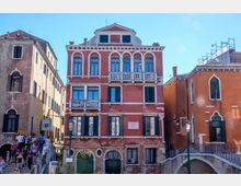 Facciata di un edificio storico veneziano in mattoni rossi, con finestre ad arco e balconi in pietra bianca, affacciato su un canale. Ai lati si vedono altri palazzi colorati e un ponte in pietra, con lampioni e un cartello “Palazzo Fortuny” in strada.