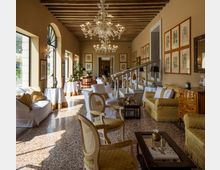 Ampio salone con soffitto a travi in legno e grandi lampadari in vetro, illuminato da alte finestre ad arco sul lato sinistro. Arredi in stile classico con divani e poltrone, tavolini, una rampa con balaustra al centro e pareti decorate da numerose cornici con stampe botaniche.