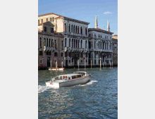 Facciate di palazzi storici veneziani affacciati su un ampio canale, con finestre ad arco e balconi in stile gotico e rinascimentale. In primo piano si vede l’acqua del canale con un motoscafo che passa, mentre sullo sfondo spiccano due sottili campanili.