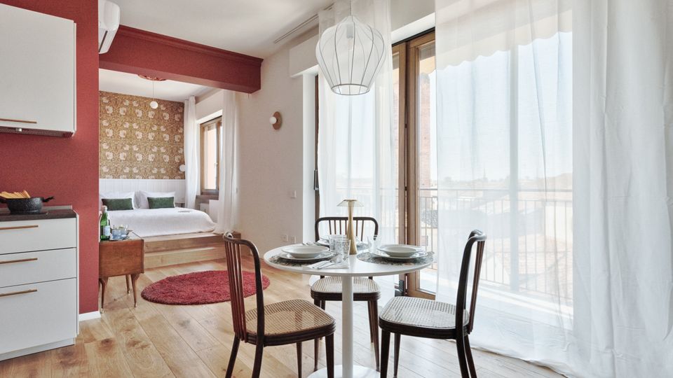Interno di un monolocale moderno con zona pranzo con tavolo rotondo e sedie, cucina a vista e grande portafinestra con tende bianche che dà su un balcone. Sul fondo si vede la zona notte rialzata con letto, parete decorata e pavimento in legno chiaro.