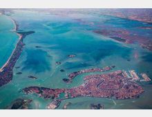 Veduta aerea di una città lagunare circondata da canali e acque turchesi, con isole minori sparse e una lunga lingua di terra costiera sul lato sinistro. Si distinguono il tessuto urbano denso, le vie d’acqua principali e aree portuali o industriali verso destra.