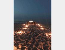 Spiaggia notturna con un tavolo e sedie posizionati sulla sabbia, circondati da piccole luci a terra disposte in cerchi. Sullo sfondo si intravede il mare scuro sotto un cielo nuvoloso.