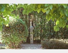 Piccolo giardino con sentiero di ghiaia, siepi potate e alberi che formano una cornice di foglie in primo piano. Al centro si vede un pilastro in mattoni con un elemento decorativo in pietra davanti a una folta siepe.