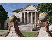 Facciata di una villa in stile neoclassico con portico a colonne e ampia scalinata centrale, immersa in un giardino. In primo piano due pilastri in mattoni e intonaco sormontati da sfere decorative incorniciano la vista dell’edificio.