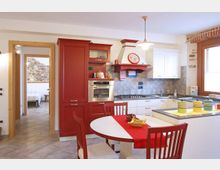 Cucina abitabile in stile rustico con mobili bianchi e rossi, forno incassato, piano cottura e cappa con mensola. In primo piano un tavolo rotondo con sedie rosse e runner, e sullo sfondo una porta verso un’altra stanza con parete in pietra e una finestra che illumina l’ambiente.