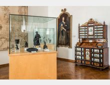 Sala museale con pavimento in parquet e pareti bianche, con una grande vetrina espositiva in vetro su base in legno contenente piccole sculture. Sulla parete sono presenti una grande mappa antica e un ritratto incorniciato, accanto a un elaborato mobile-scrivania intarsiato con numerosi cassetti decorati.