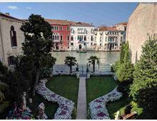 Giardino formale lungo un canale, con vialetto centrale che conduce a un cancello sul bordo dell’acqua, aiuole fiorite simmetriche e palme. Sullo sfondo si vedono facciate storiche colorate affacciate sul canale e muri laterali che incorniciano la vista.