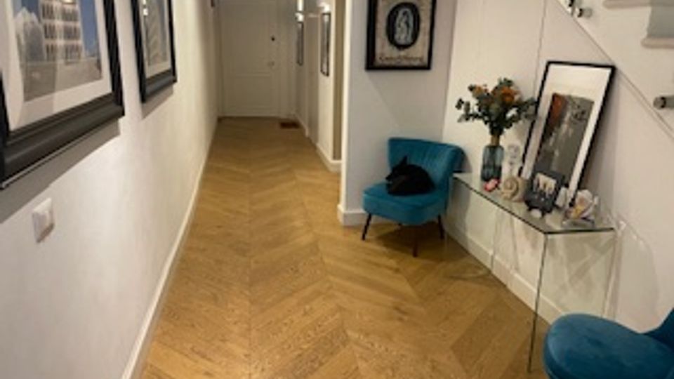 Corridoio interno di un’abitazione con pavimento in parquet a spina di pesce e pareti bianche, decorato con quadri incorniciati. A destra ci sono una consolle con specchio, oggetti e fiori, due sedie imbottite turchesi e una scala con gradini bianchi.
