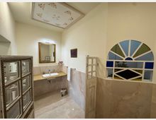 Bagno luminoso con pareti e pavimento in piastrelle chiare, lavandino con piano in pietra e grande specchio rettangolare sopra. A destra c’è una finestra ad arco con riquadri geometrici colorati e un soffitto alto con riquadro decorato.