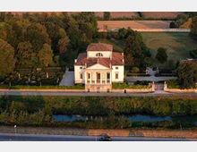 Villa storica di campagna con facciata neoclassica e portico a colonne, circondata da alberi e campi coltivati. In primo piano si vedono una strada e un canale d’acqua parallelo al viale d’ingresso con cancello.